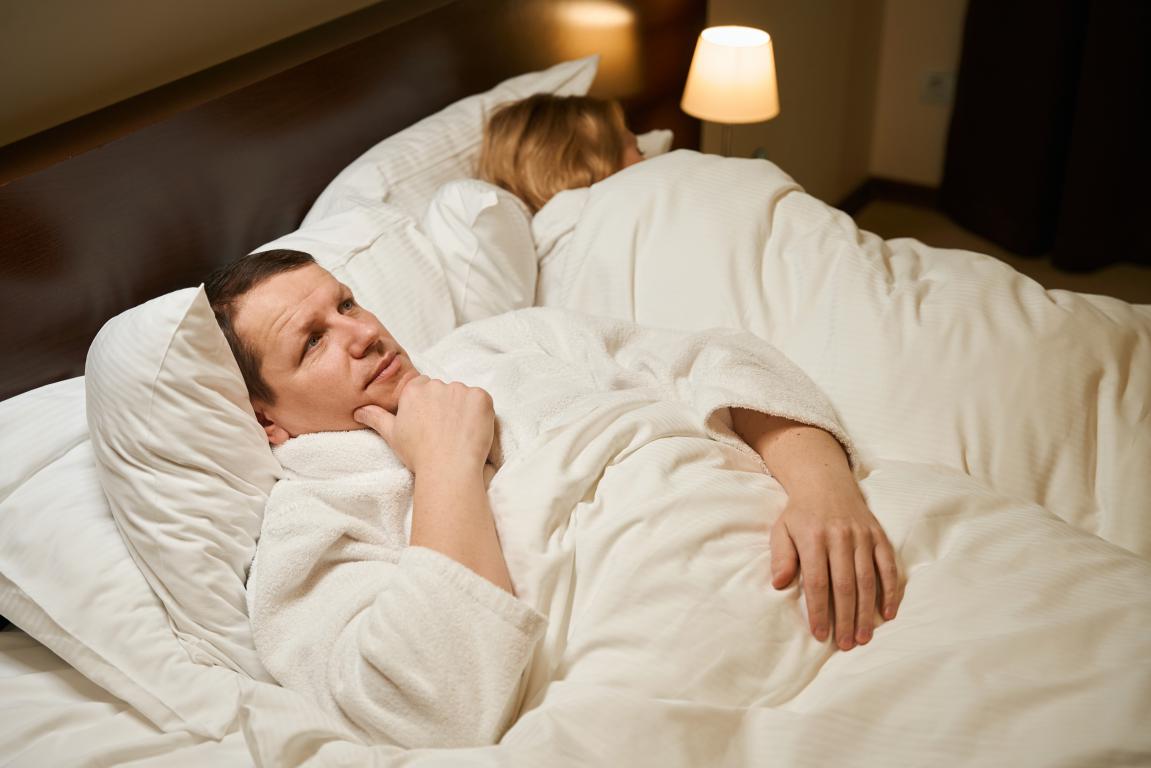 A man in a white bathrobe lies awake in bed, staring at the ceiling with a thoughtful or exasperated expression, due to a partner's snoring,