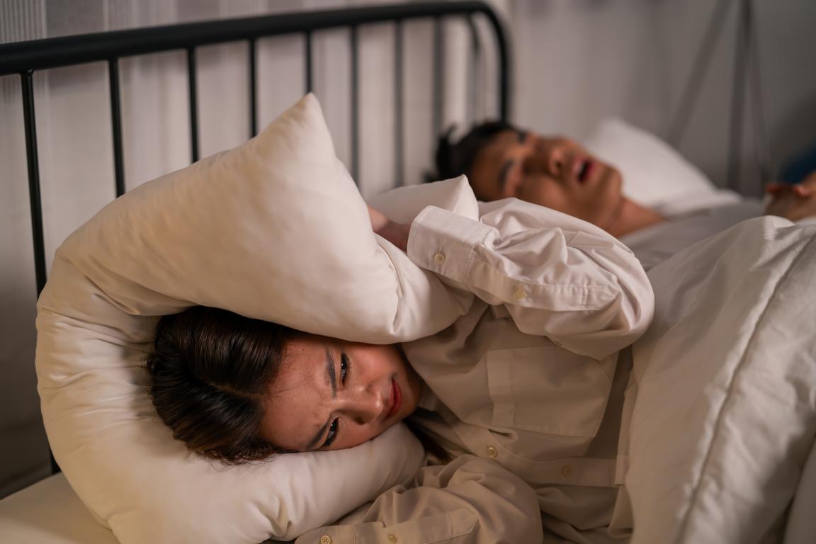 A woman lies in bed with her head buried under a pillow, grimacing, attempting to muffle the sound of a man snoring loudly behind her