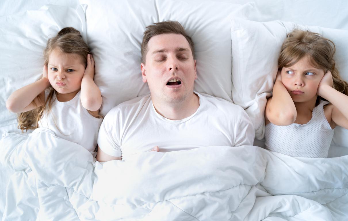 A man sleeps on his back with his mouth open, snoring. Two young girls on either side of him cover their ears with annoyed expressions, unable to sleep due to the noise.