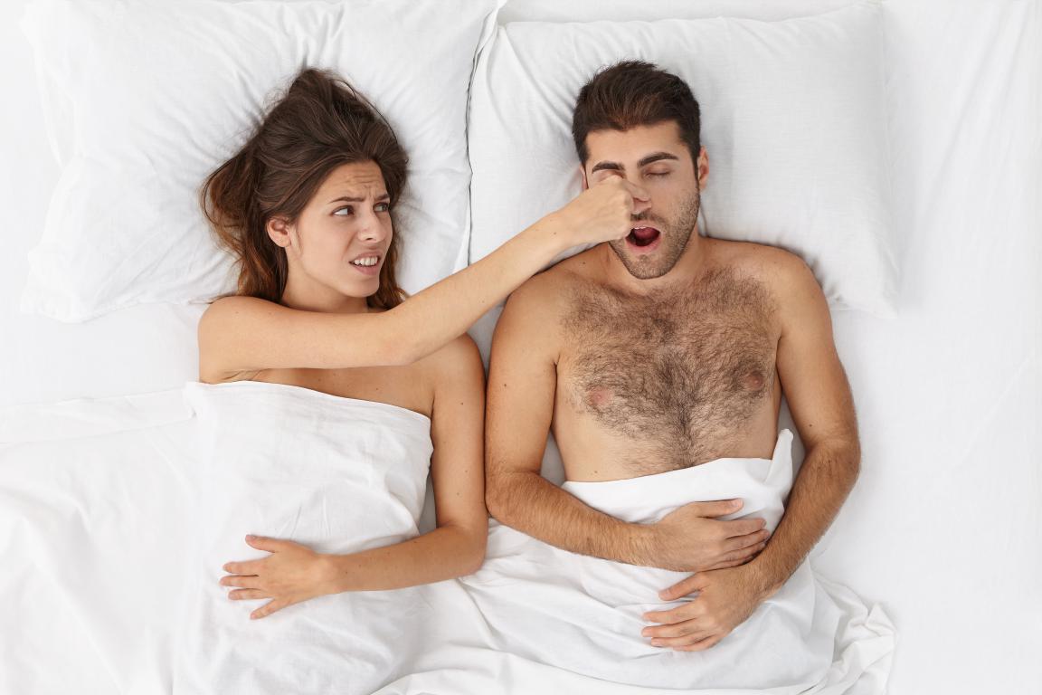 From an overhead view, a woman looks disgusted as she pinches the nose of a loudly snoring, shirtless man lying next to her in a white bed.