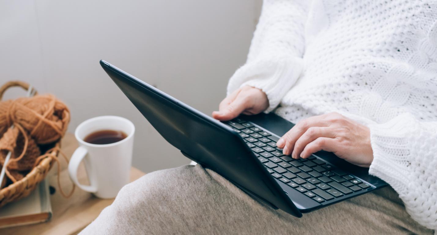 A person in a white knitted jumper and light trousers sitting, typing on a dark laptop. A white mug with a hot drink and a basket of yarn are on a table next to them.