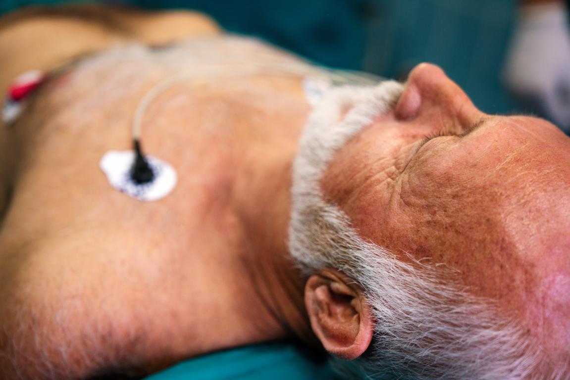  Close-up of an older man with a white beard lying down, with medical electrodes attached to his chest.