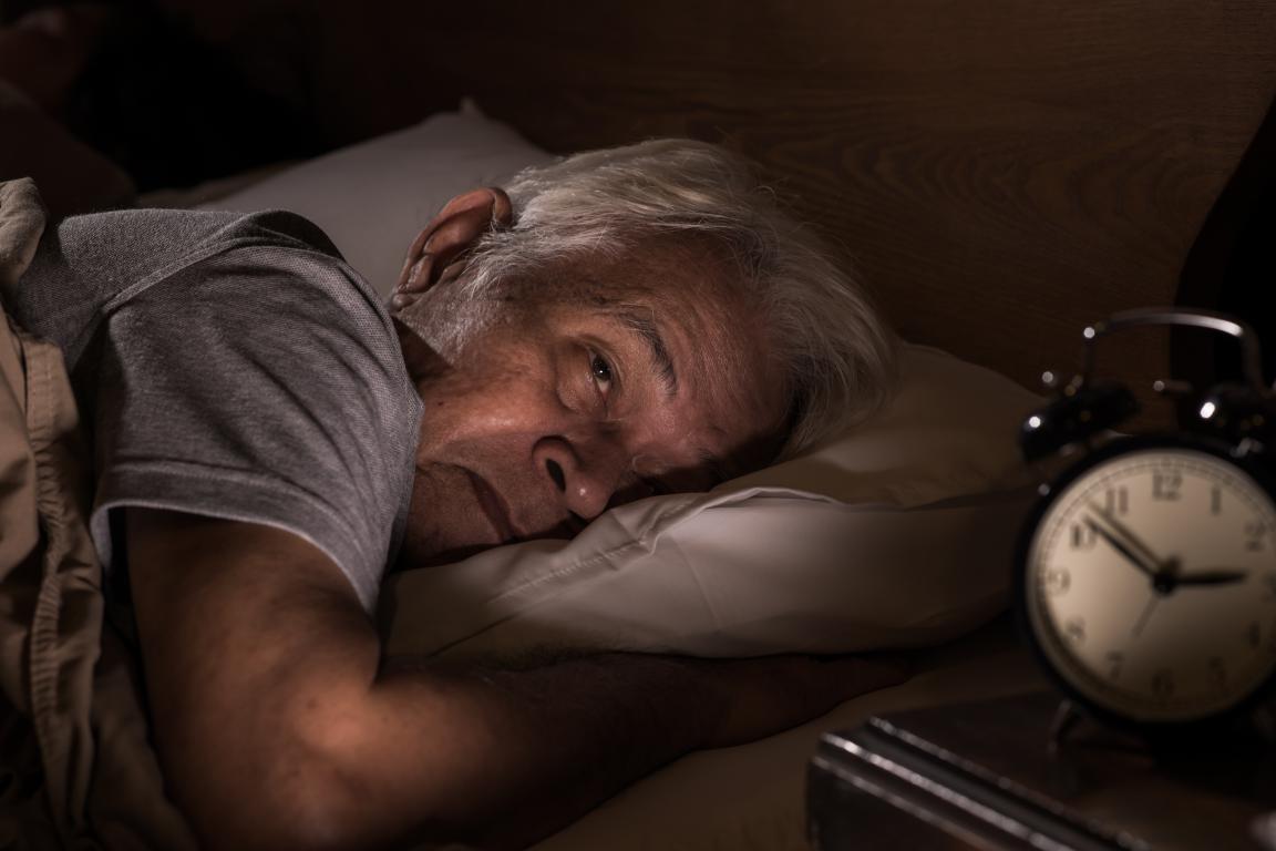 An elderly man lies awake in bed with an alarm clock nearby, illustrating the common symptom of fragmented sleep often associated with a high AHI.