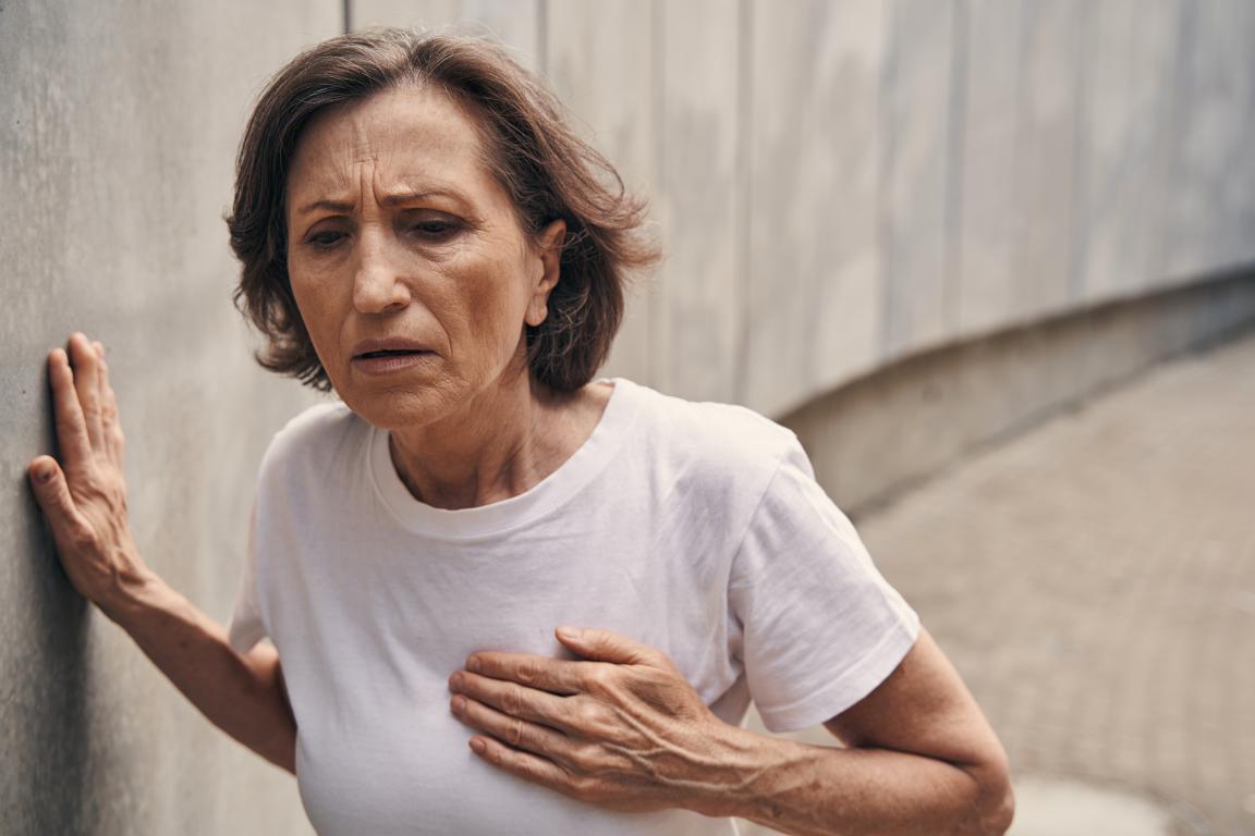 An older woman looks distressed, clutching her chest and leaning against a wall, depicting the sudden and alarming feeling of a potential heart event or ICD shock.