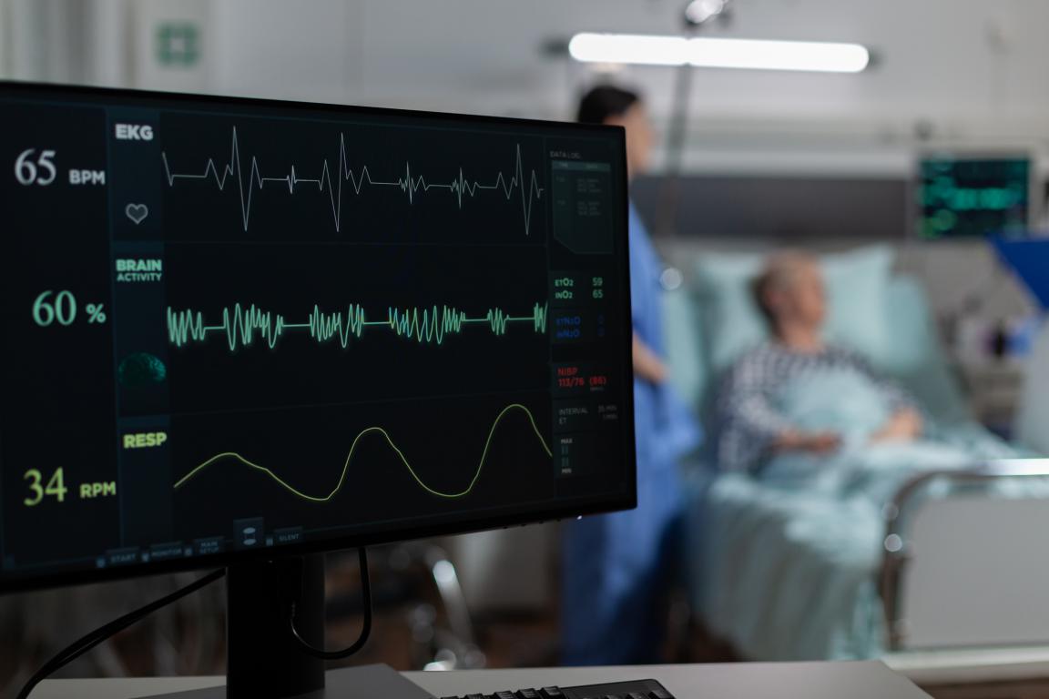 A close-up of a medical monitor displaying an EKG waveform, brain activity, and vital signs, with a patient and nurse blurred in the background