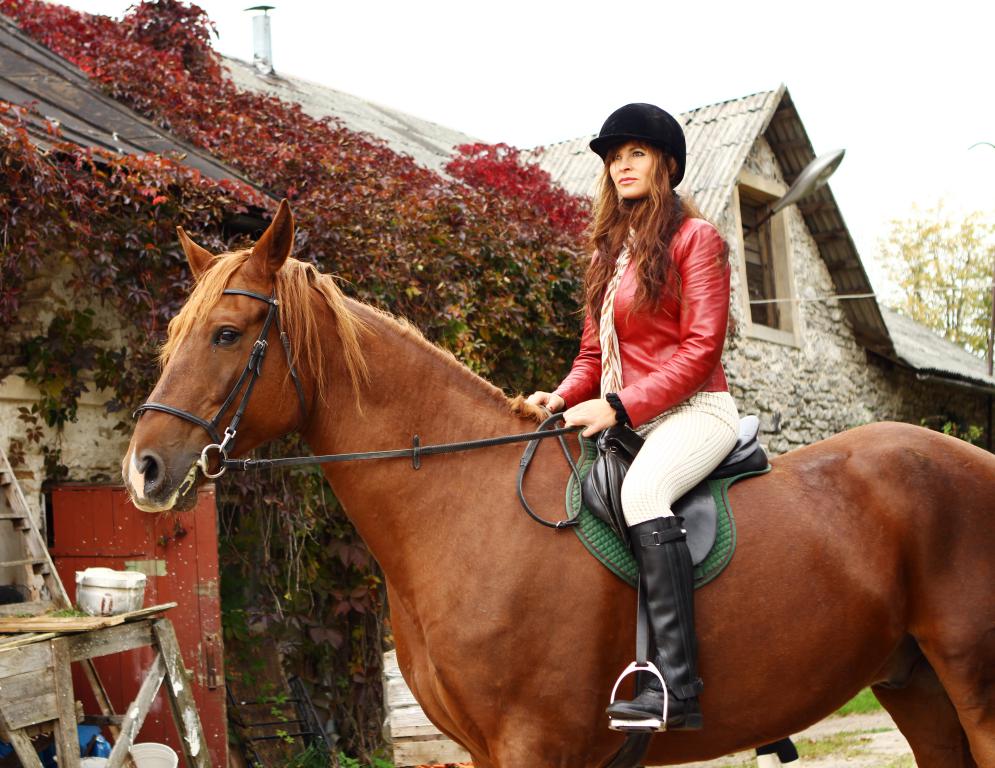  A female rider wearing a helmet, red leather jacket, and riding boots sits atop a chestnut horse, with an old stone building in the background.