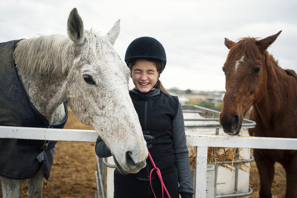A smiling young girl in a riding helmet stands between a grey horse wearing a rug and a brown horse in a field, showing a bond between child and equines.
