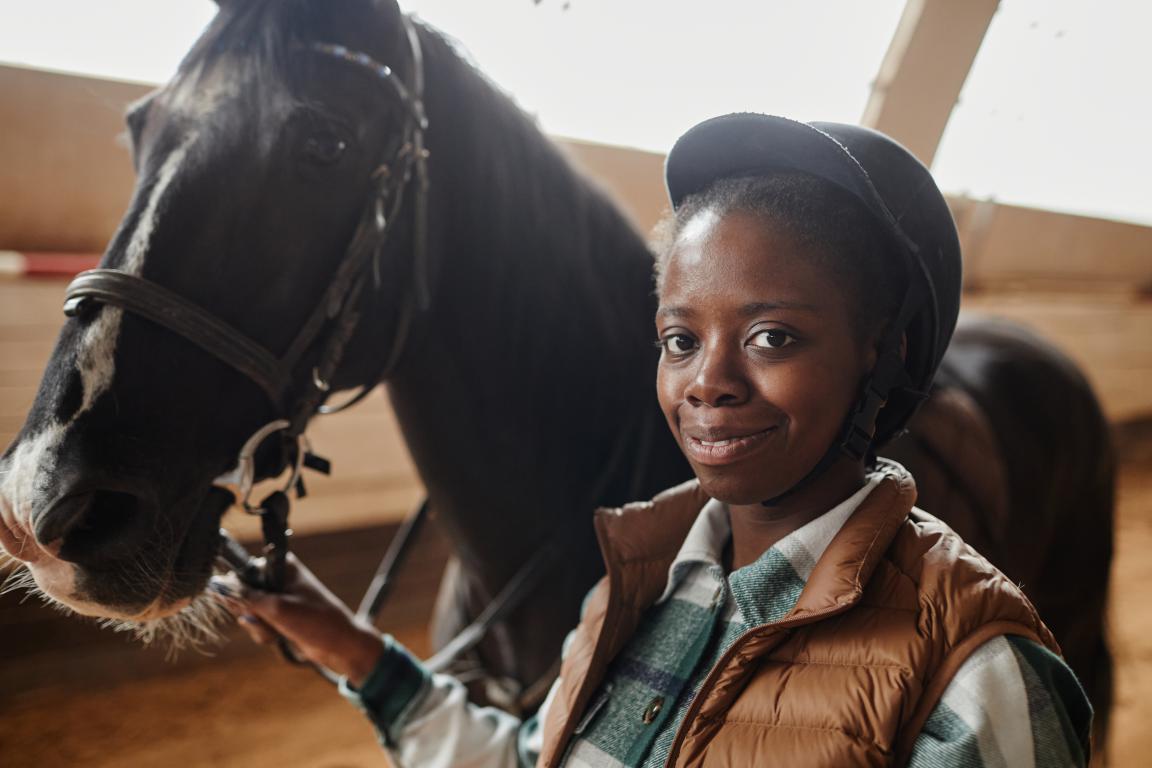 A smiling young Black woman wearing a riding helmet and a puffer vest holds the bridle of a dark bay horse in an indoor arena, looking at the camera.