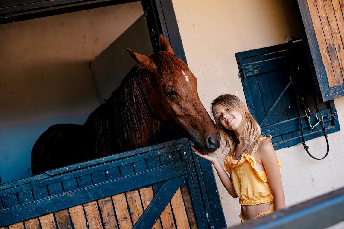 A young girl in a yellow top stands smiling next to a stable door, reaching out to touch the muzzle of a friendly brown horse looking out from its stall.