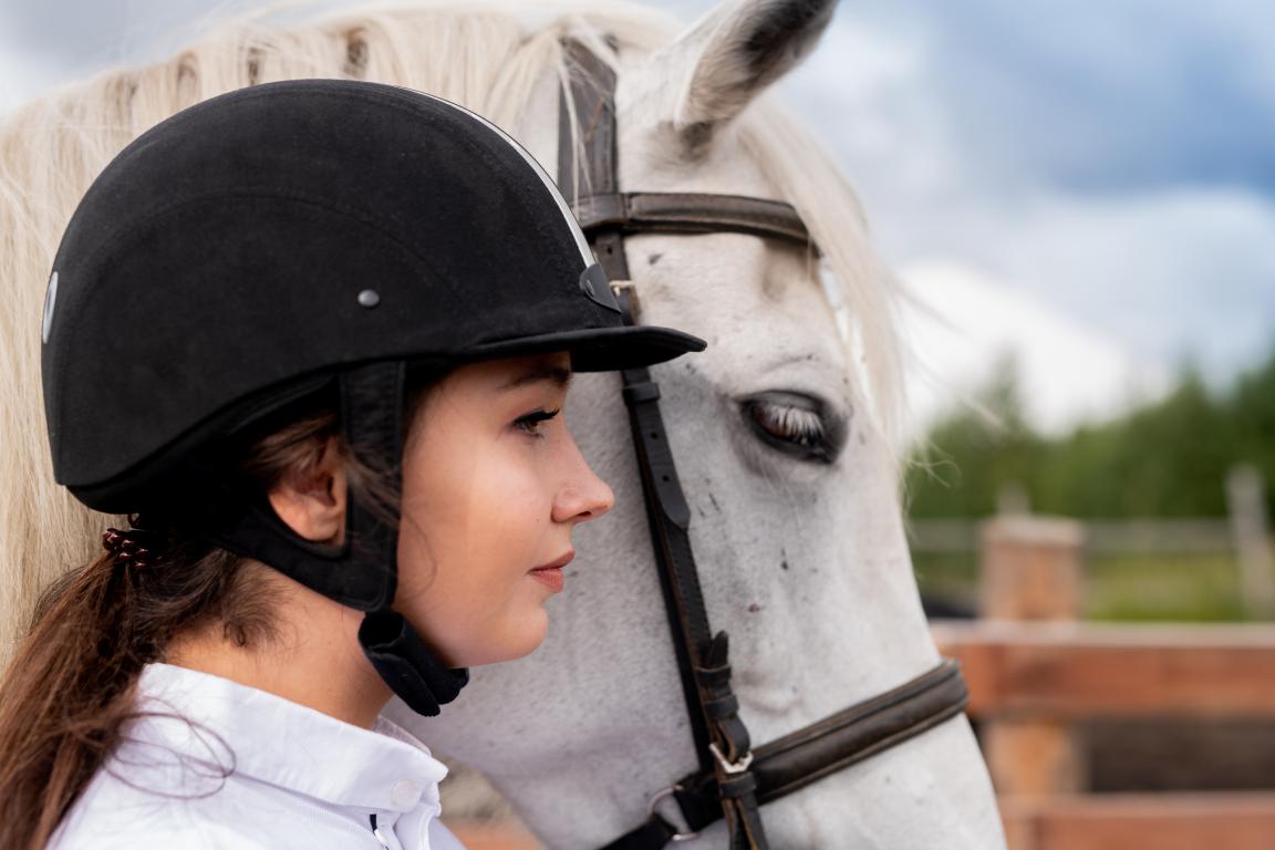 Close-up profile of a young woman in a riding helmet next to a grey horse, highlighting the connection between owner and animal.
