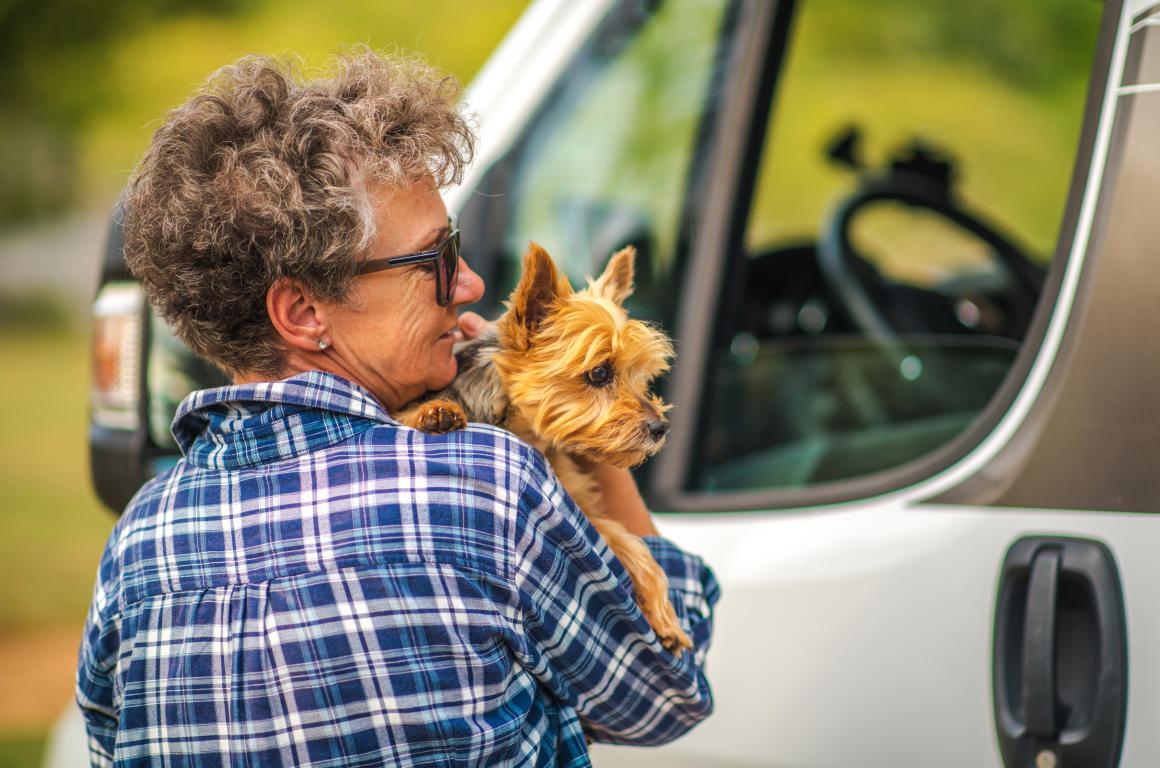 An older woman in a plaid shirt and sunglasses holds a small, scruffy terrier-mix dog close to her, with the side of a white vehicle visible behind them. The dog, likely an assistance dog, looks attentive and comfortable in the woman's arms