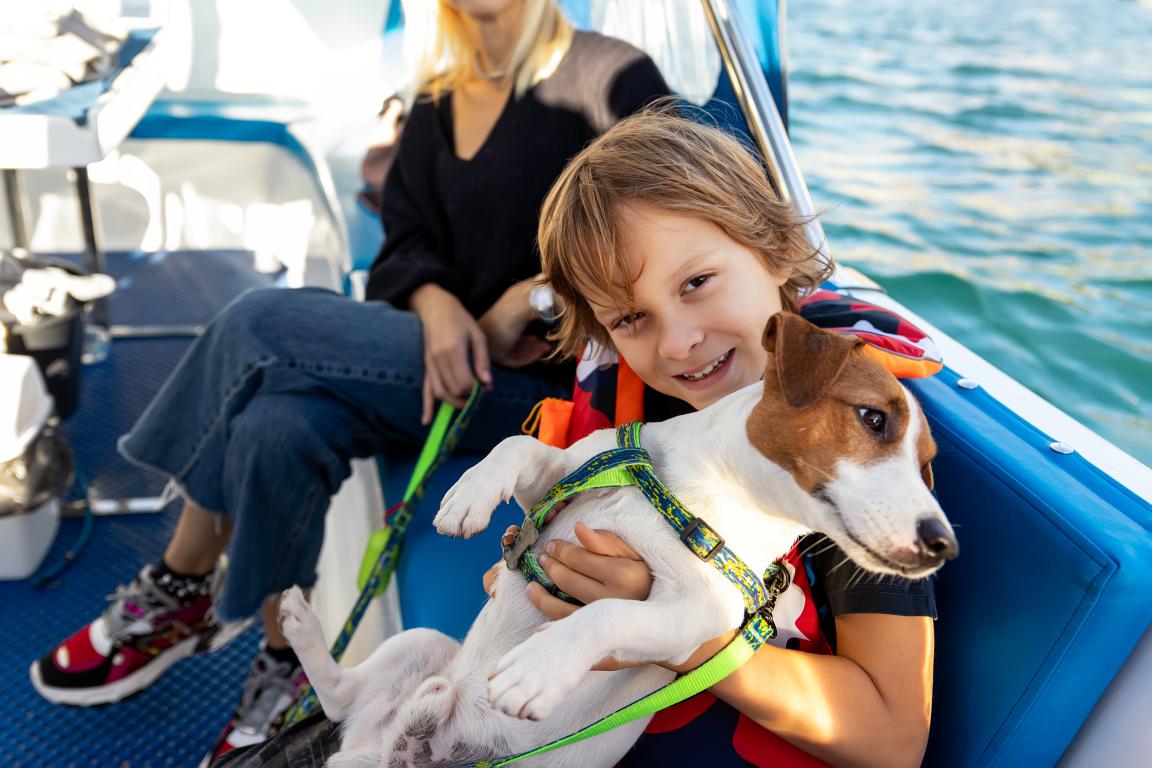 A smiling child holds a small, harnessed assistance dog on a boat, with calm water visible in the background