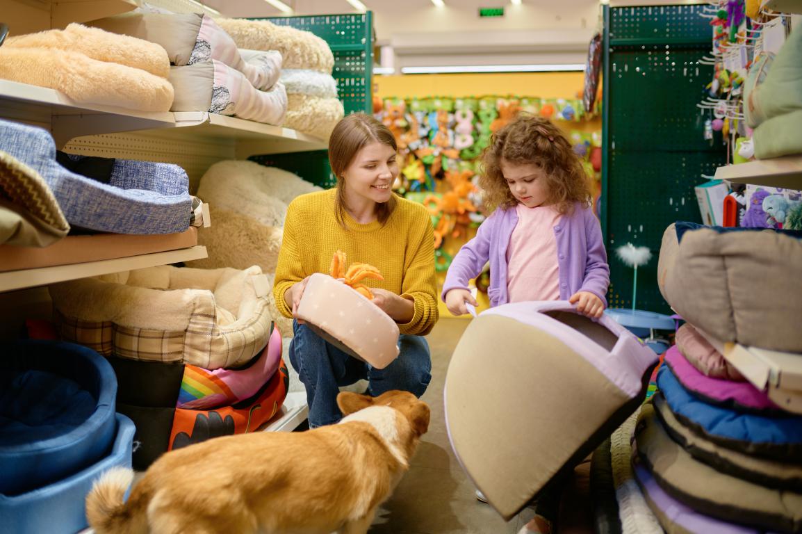 A woman and a young girl are squatting down in a pet supply store aisle, looking at pet beds with a small, brown and white dog (possibly a Corgi mix) standing in front of them. The woman holds a pink pet bed, and the girl points at another.
