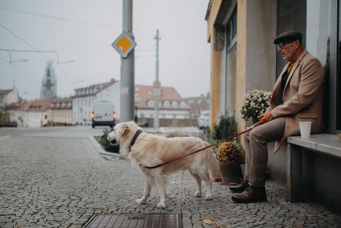 An older man wearing a hat and sunglasses, dressed in a beige suit, sits on a bench on a cobblestone street, with a light-colored Golden Retriever on a red leash standing attentively beside him. Buildings and blurred street scenes are in the background.