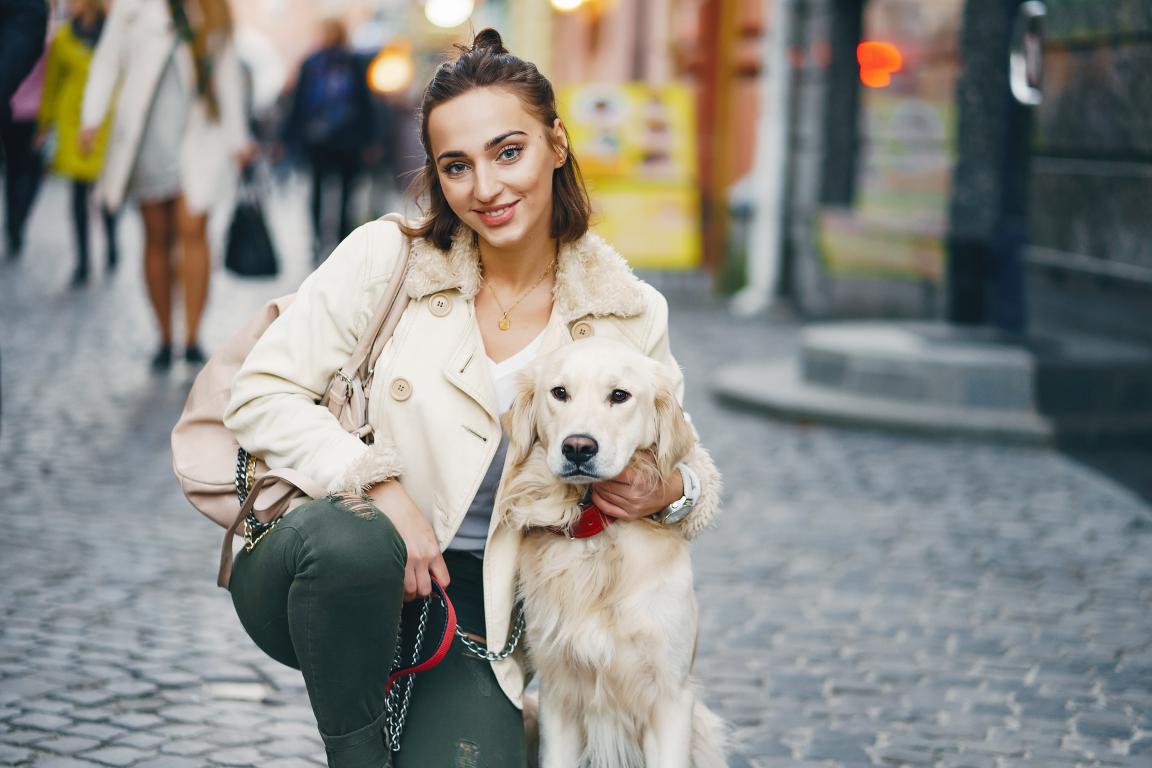 A smiling young woman with dark hair, wearing a cream coat and green trousers, crouches down on a cobblestone street, holding a leash attached to a cream-colored Golden Retriever. The dog looks calmly at the camera, and blurred city life is in the background.