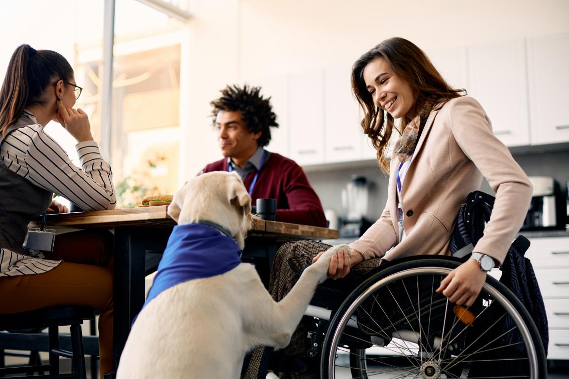 A woman in a wheelchair smiles as she gently holds the paw of a cream-colored Labrador assistance dog wearing a blue bandana. They are at a table in what appears to be an office or modern kitchen setting, with two other people blurred in the background.