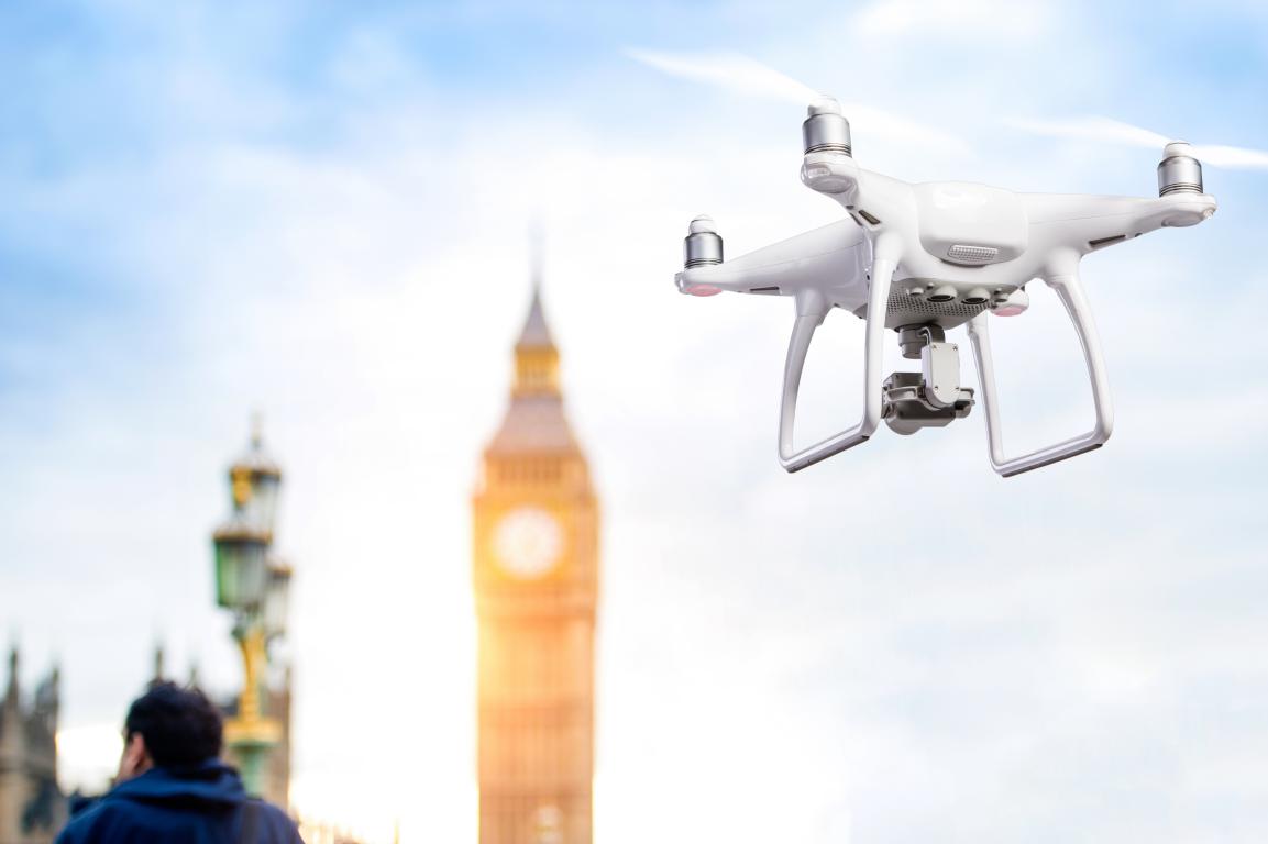 A white drone flying in the sky with Big Ben blurred in the background, a person looking up towards the drone