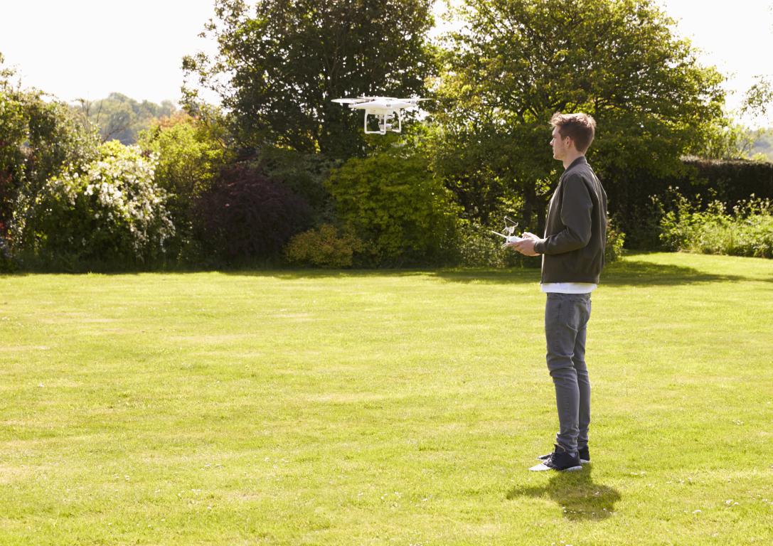 A young man standing in a large green garden, operating a white drone that is hovering in the air