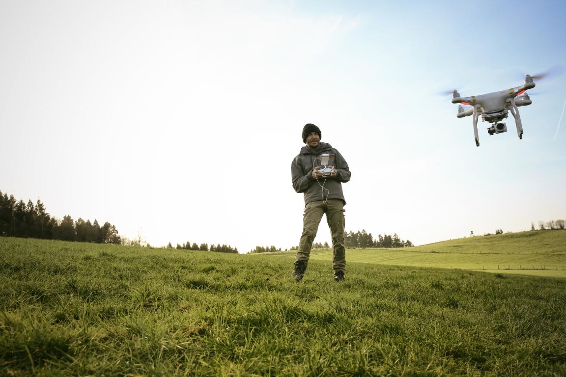  A person holding a drone controller in a grassy field, with a white drone flying in the distance against a bright sky