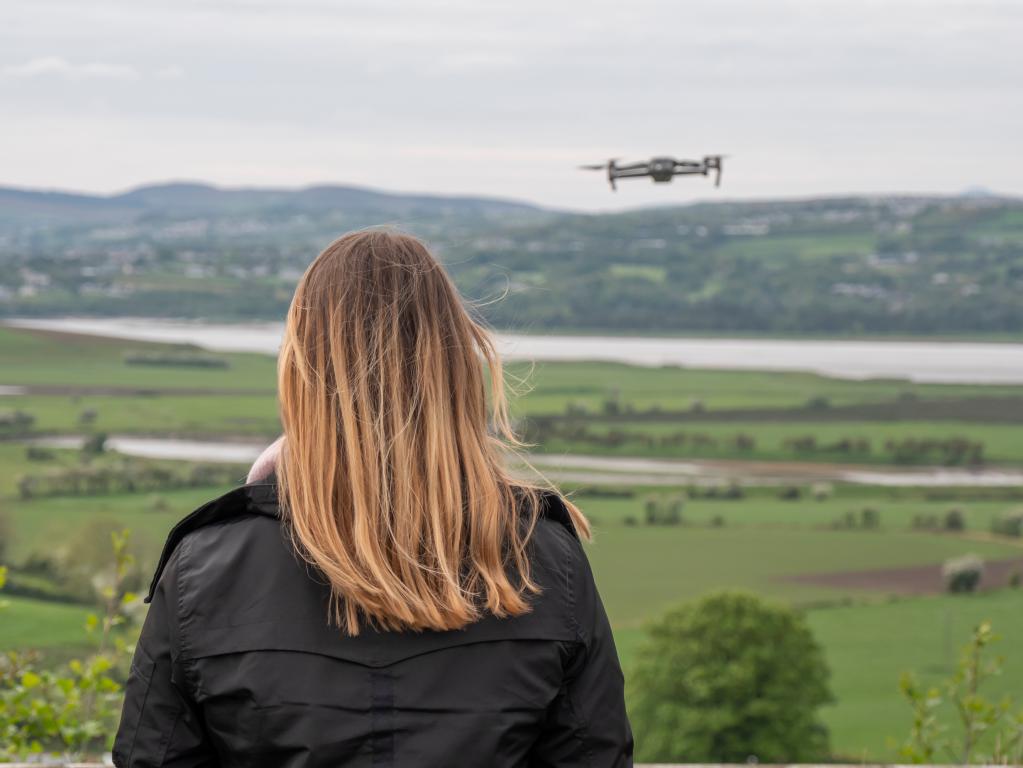 A woman with long hair seen from behind, looking out over a green landscape with a drone flying in the distance