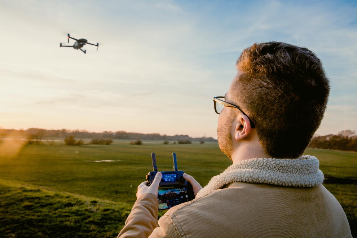 A man seen from over his shoulder, wearing a sherpa-lined jacket and glasses, operating a drone with a controller, looking at a drone flying in a field at sunset