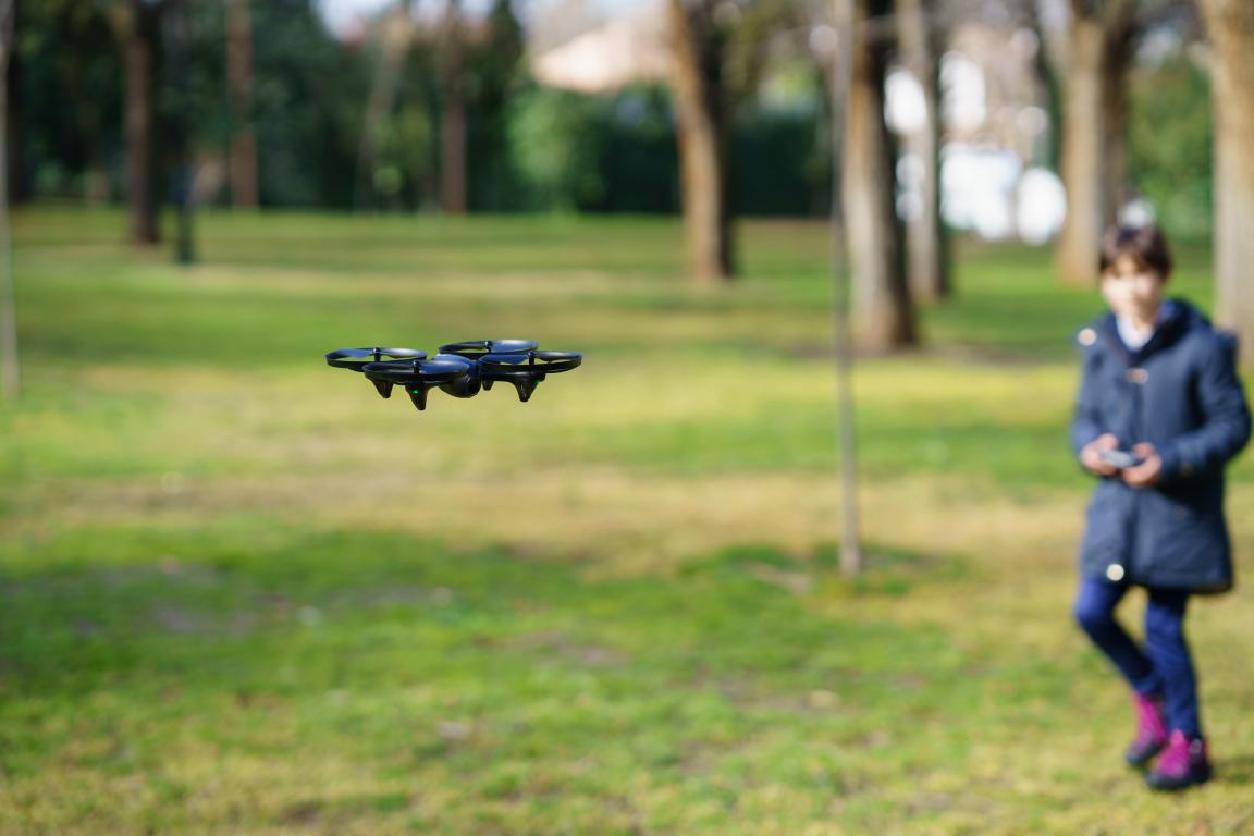 A small black drone hovering in a park, with a young child blurred in the background looking towards the drone