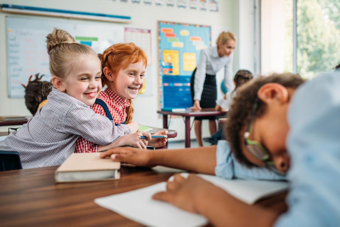 Two happy primary school girls smiling at their desk while a teacher supervises the classroom in the background.