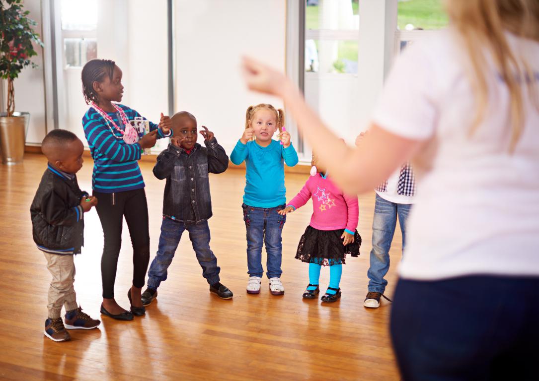 A group of diverse young primary school children listening to their teacher during a group activity in a school hall.