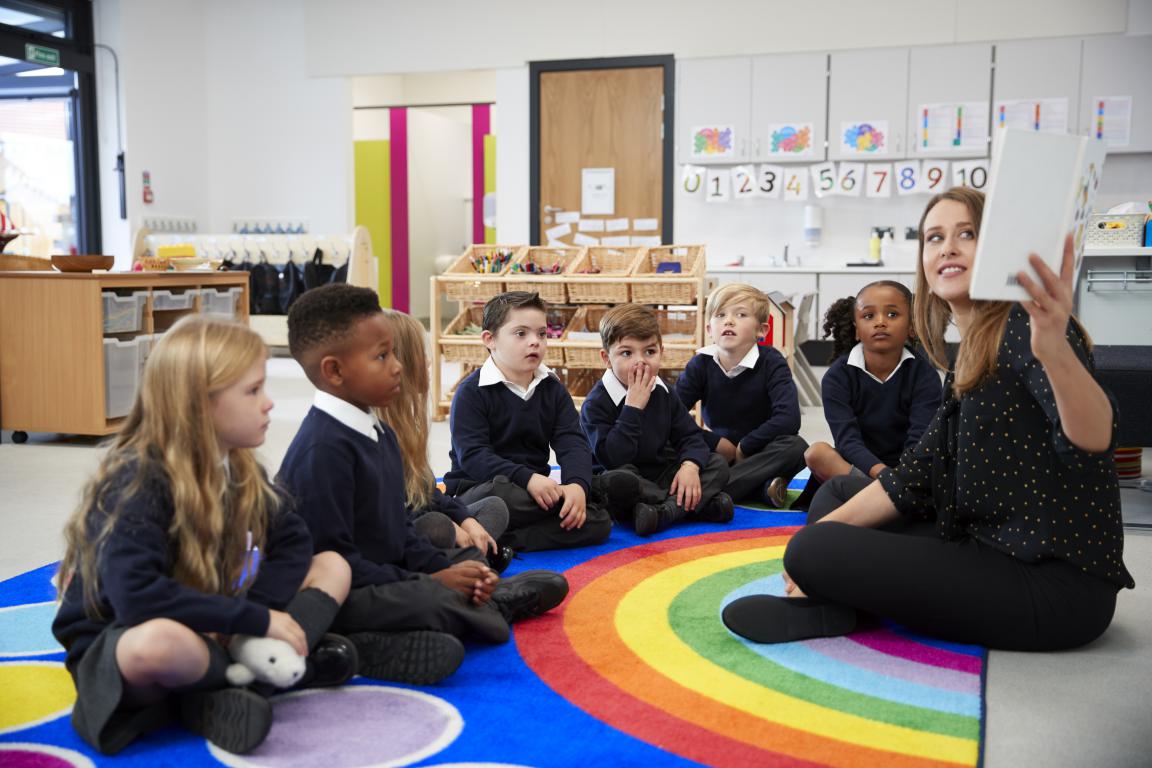 A teacher reading a story to a group of young primary school children sitting together on a colourful classroom carpet.