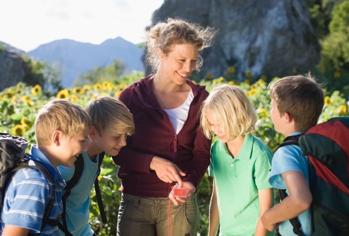 A female teacher smiles as she shows a group of four primary school children how to use a compass in a field of sunflowers.