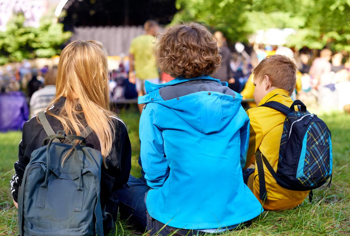 The backs of three children with backpacks as they sit on the grass, watching an event during a school trip.