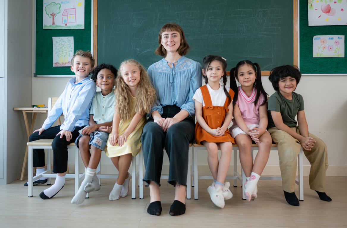 A female teacher sits on a chair in her classroom, surrounded by a smiling and diverse group of primary school pupils.
