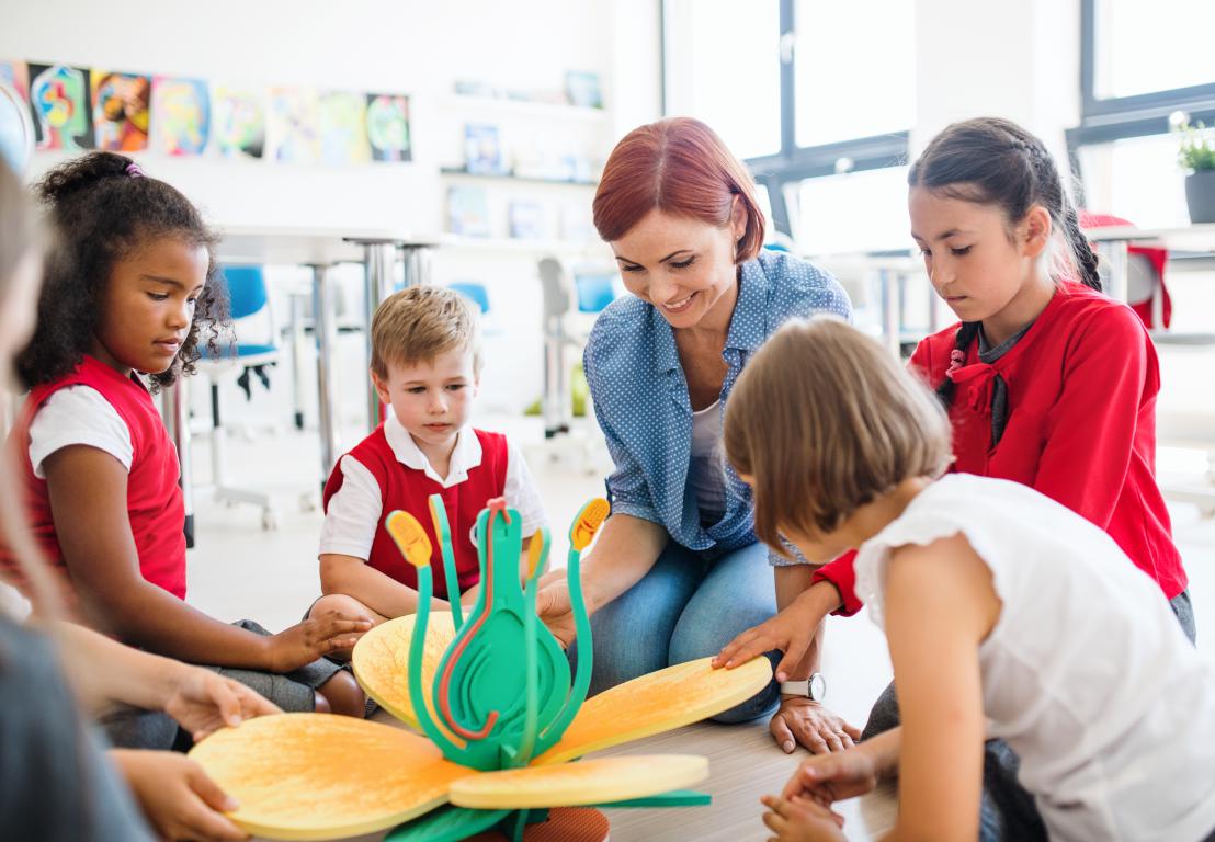 A female teacher with red hair sits on the classroom floor with a group of young, diverse pupils, looking at a large flower model.