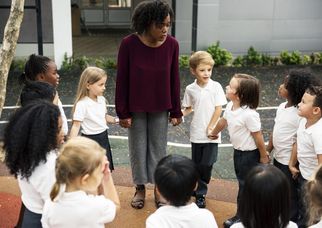 A Black female teacher stands in a circle holding hands with a group of young primary school pupils in the playground.