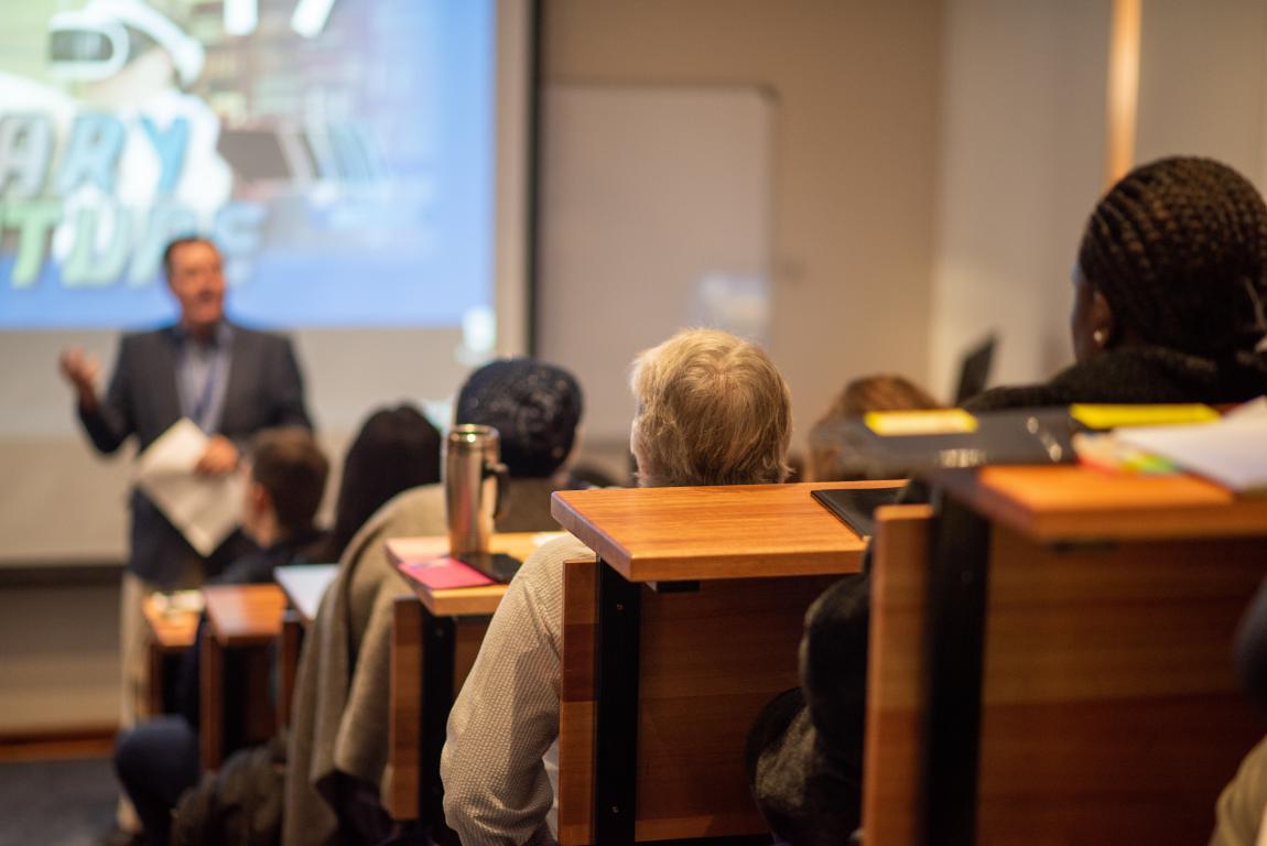  Students attending a lecture in a tiered lecture theatre, focusing on a speaker and presentation screen.