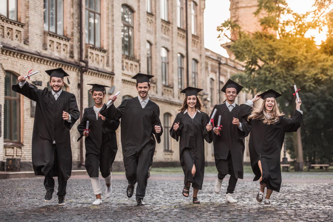 A diverse group of six graduates running excitedly on a path away from a historic university building while wearing their caps and gowns