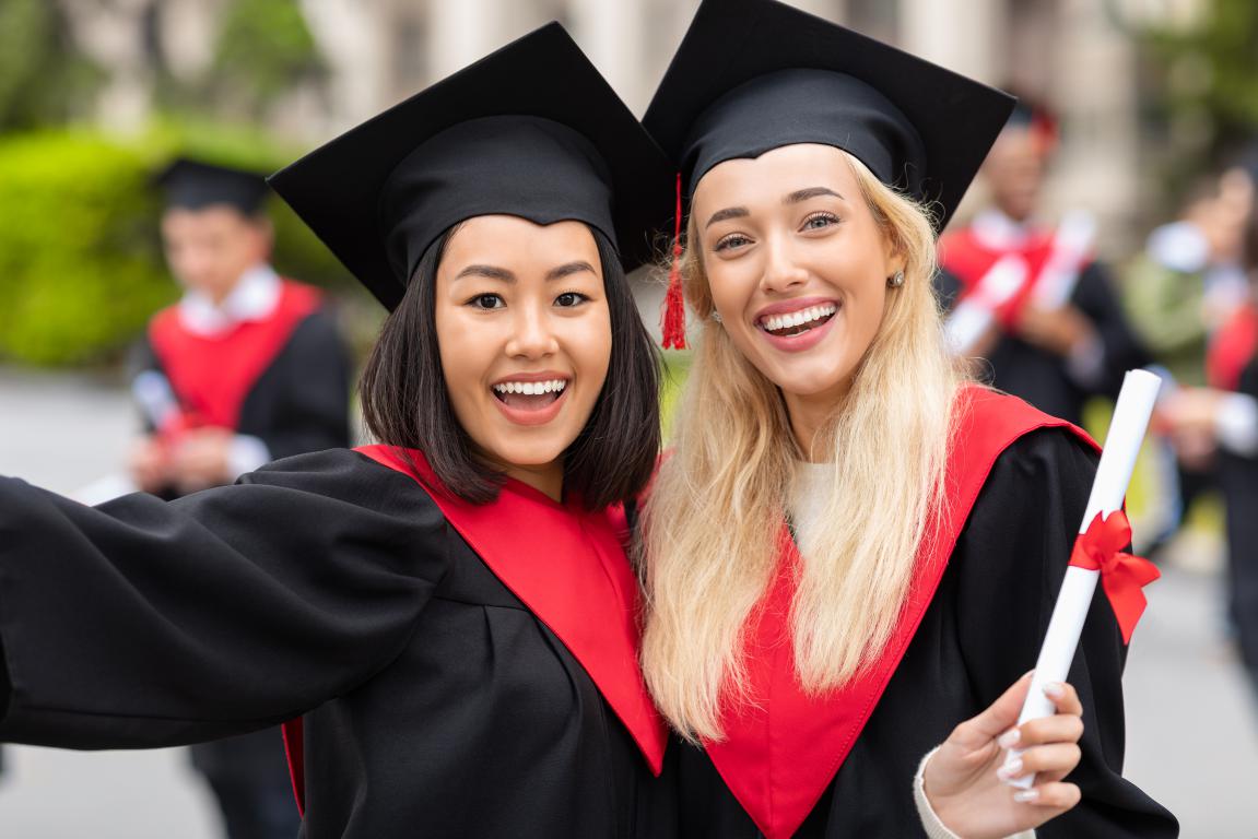 Two happy students in graduation gowns and caps taking a selfie, holding their diplomas