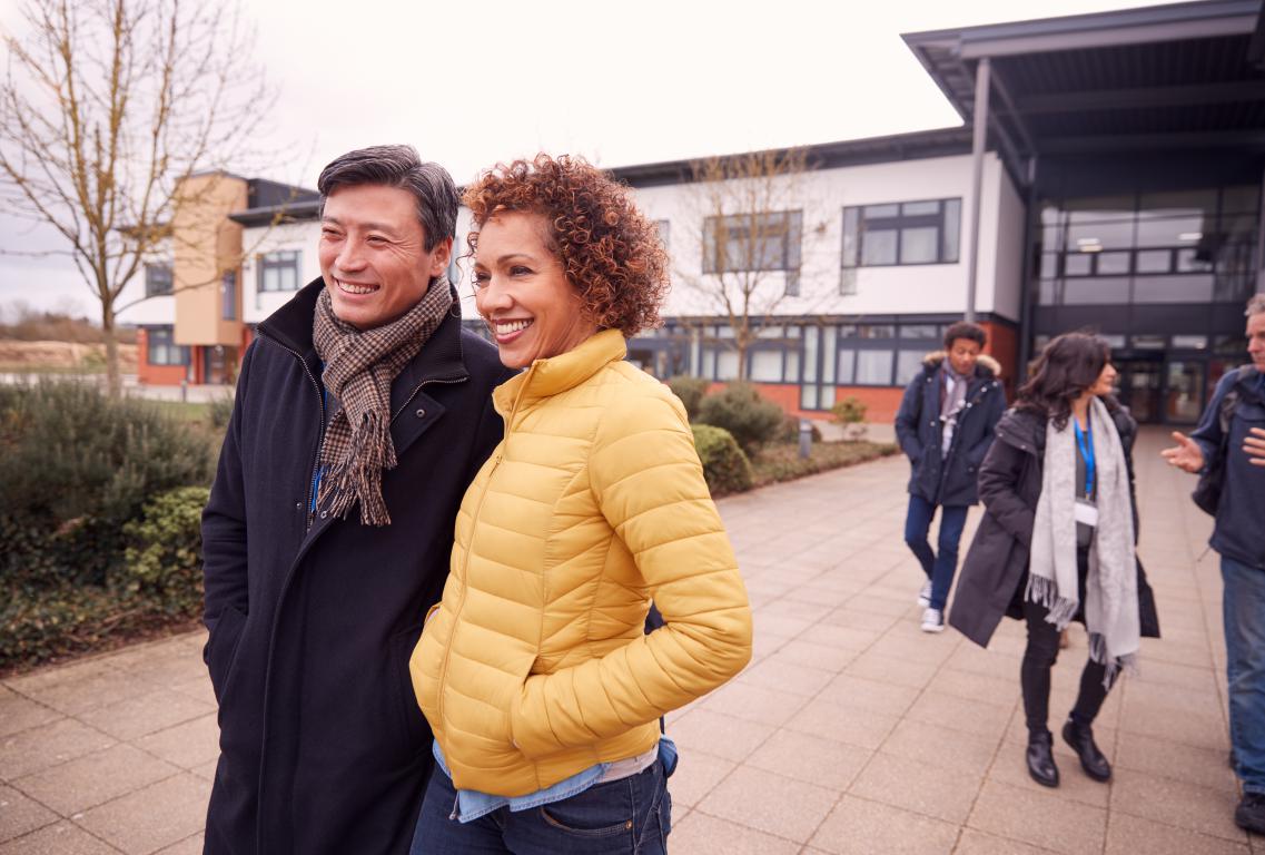 A smiling male and female staff member or mature students talk outside a modern university administrative building.