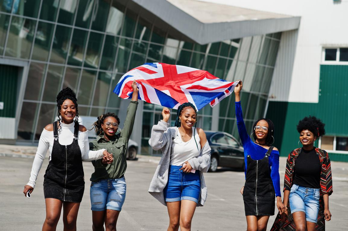 A diverse group of five female students smiling and holding up a large Union Jack flag outdoors on campus
