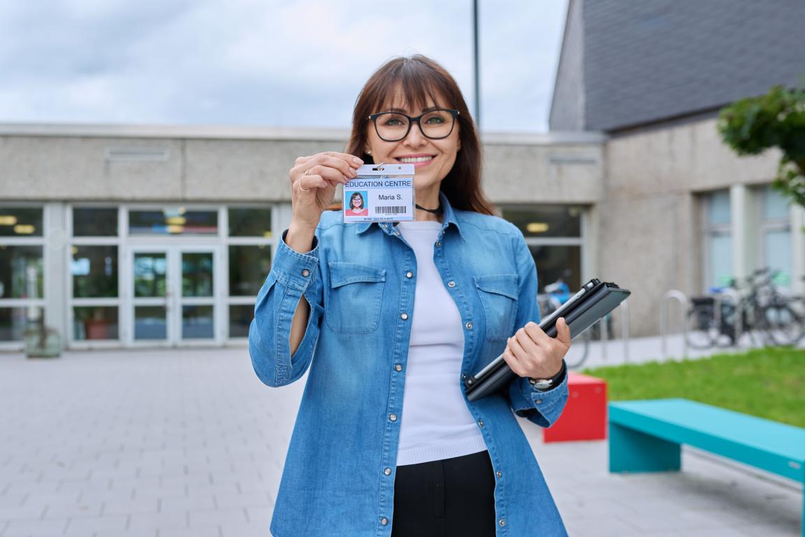 A woman (staff or student) smiling and holding up a university identification card with her photo on it, standing in front of a modern campus building.