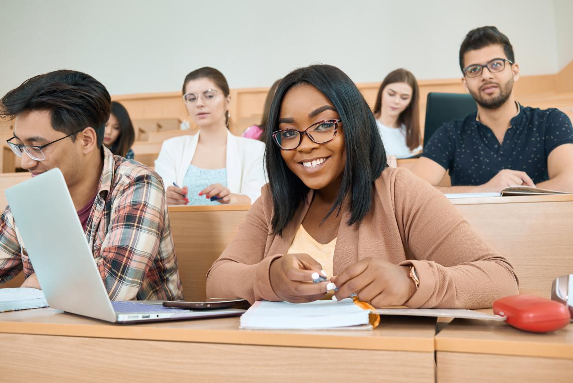 Two students studying together in the foreground of a large, tiered university lecture theatre, with other students visible working on laptops.