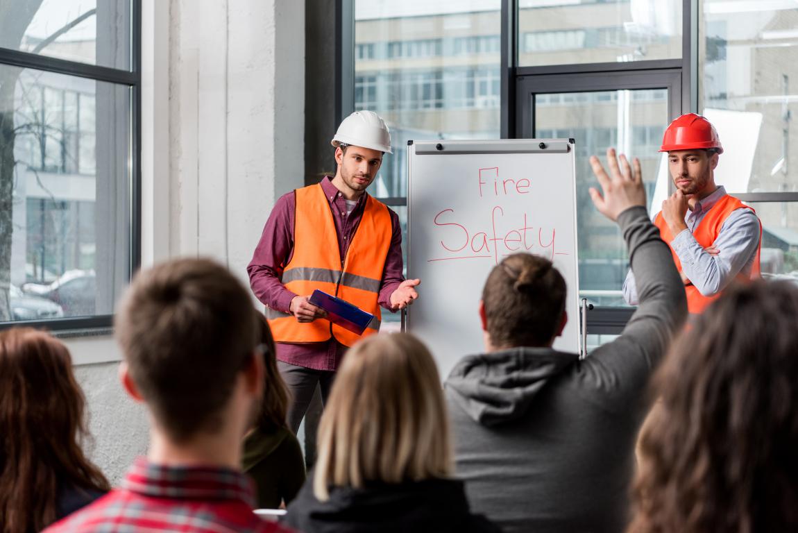 Two trainers in vests and hard hats lead a fire safety session for workers in a bright meeting room.