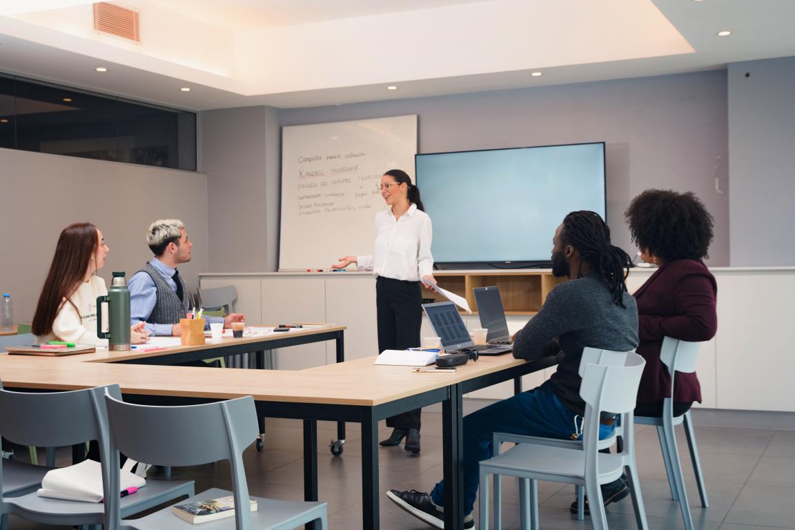 Diverse young professionals collaborate around a U-shaped table in a corporate training workshop, led by a standing female instructor at a whiteboard.
