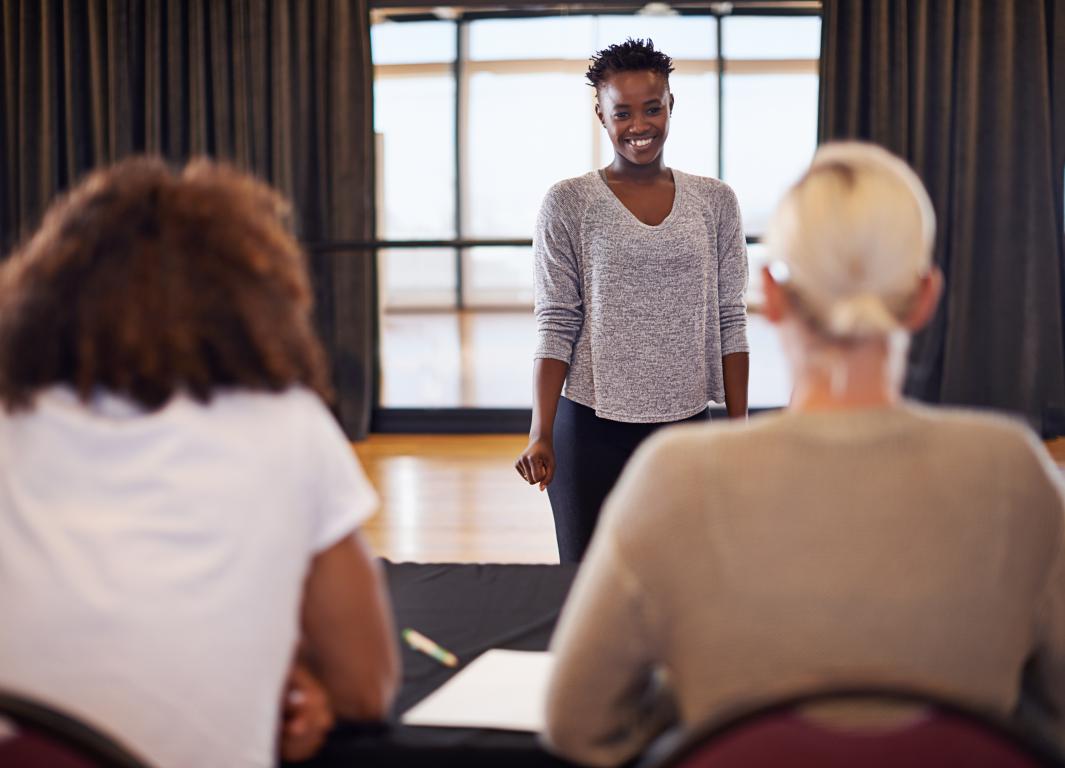 Young woman confidently leads public-speaking training, smiling at small seated group in sunlit room.