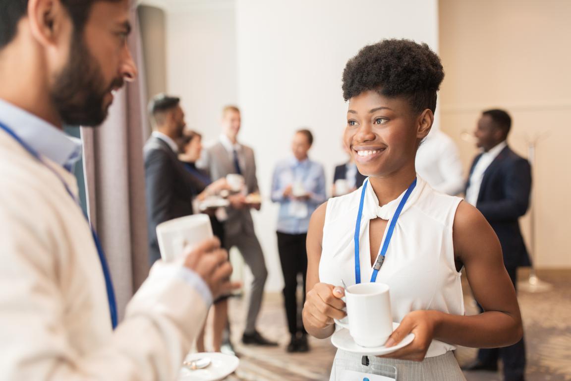 Smiling young woman wearing a blue lanyard with her training ID card visible while chatting over coffee at a professional networking event