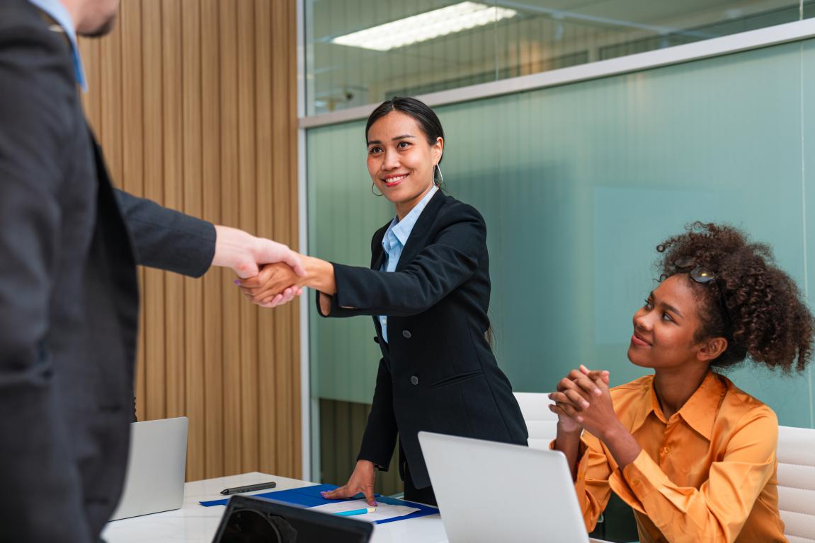 Young woman in a suit shaking hands across the interview table after confidently presenting her training ID card and qualifications