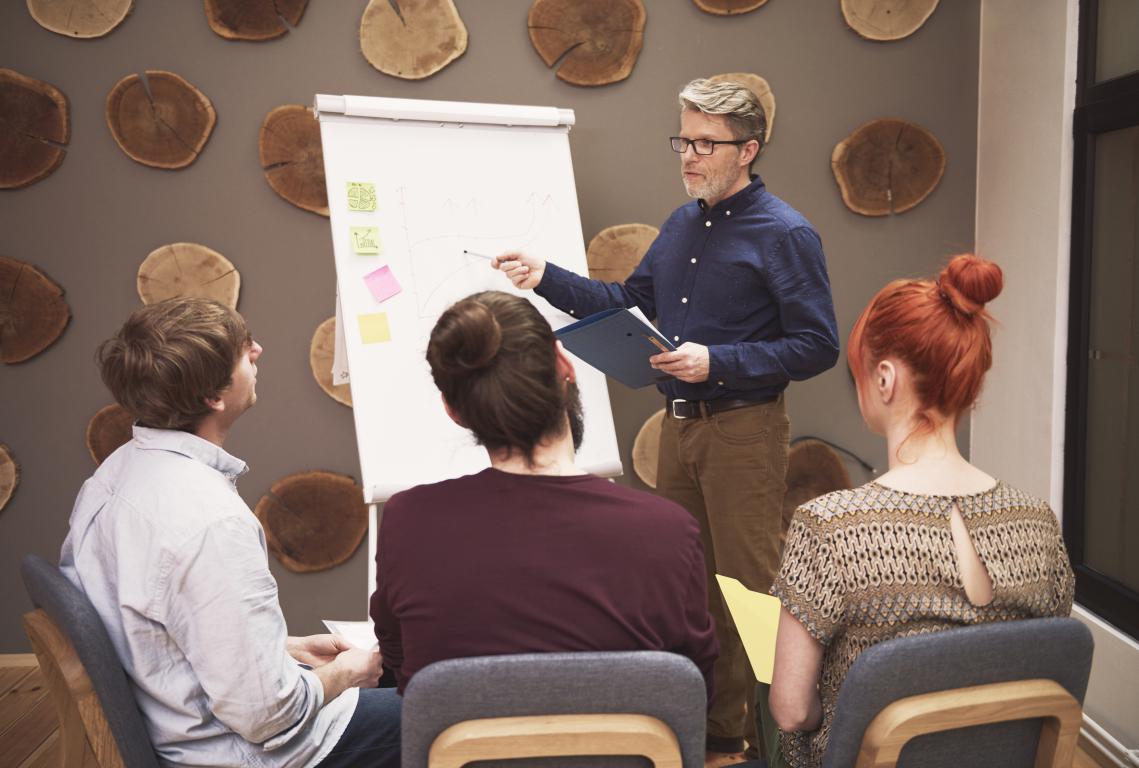 Trainer pointing at a flipchart while teaching a small group – the kind of course where a physical ID card becomes a proud takeaway.